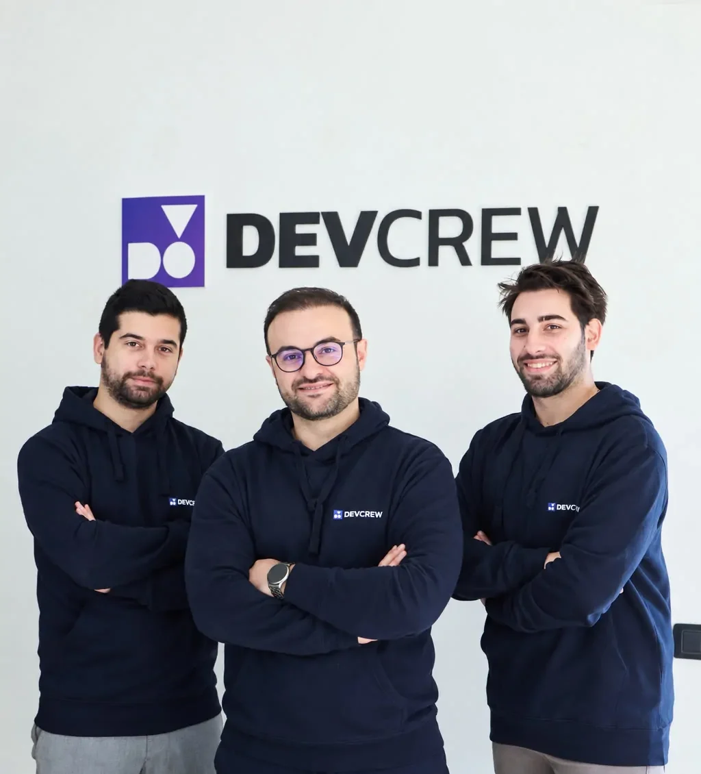 Il team DevCrew: tre sviluppatori con felpa DevCrew davanti al logo in ufficio